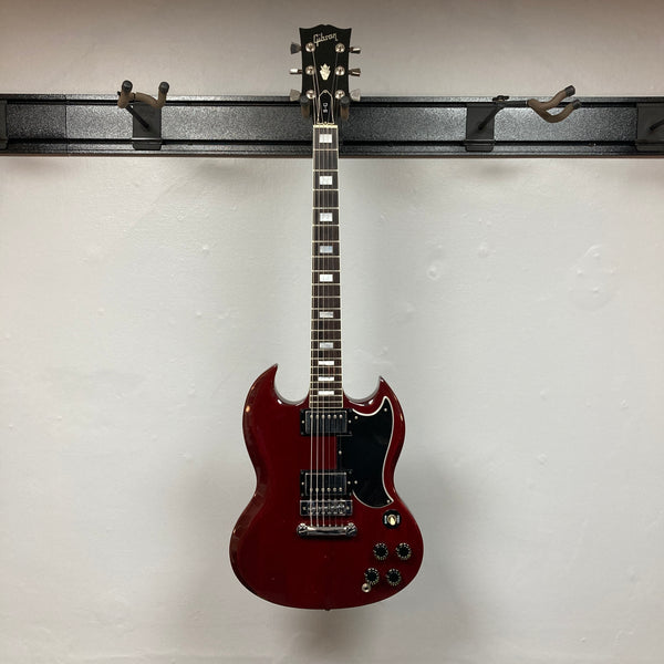 A 1977 Gibson SG Standard Cherry electric guitar by Gibson, featuring a classic double-cutaway mahogany body, two humbucker pickups, four control knobs, and a black pickguard evocative of the iconic Kalamazoo era, hangs elegantly against a plain white wall.