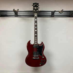 A 1977 Gibson SG Standard Cherry electric guitar by Gibson, featuring a mahogany body, black pickguard, and chrome hardware, hangs on a wall-mounted guitar holder against a white wall.