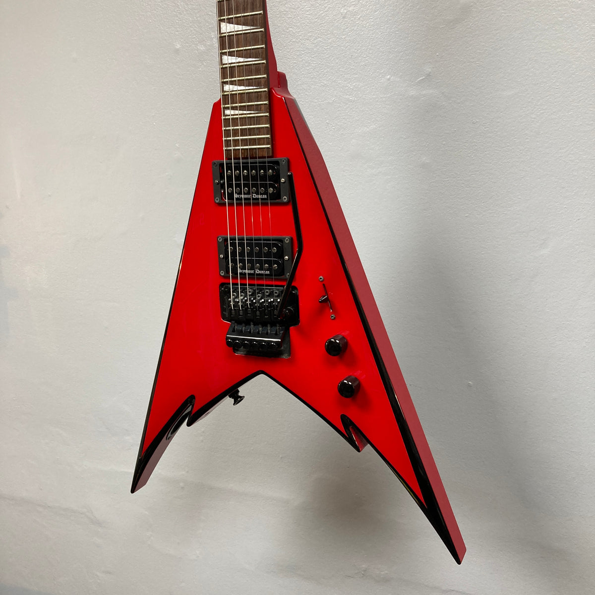 A Jackson Phil Demmel Demmelition King V guitar in striking red with a V-shaped body, featuring black hardware, dual humbucker pickups, a Floyd Rose tremolo system, and two control knobs, displayed against a plain white wall.