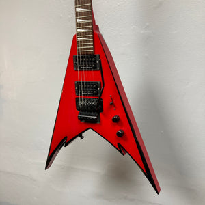 A Jackson Phil Demmel Demmelition King V guitar in striking red with a V-shaped body, featuring black hardware, dual humbucker pickups, a Floyd Rose tremolo system, and two control knobs, displayed against a plain white wall.