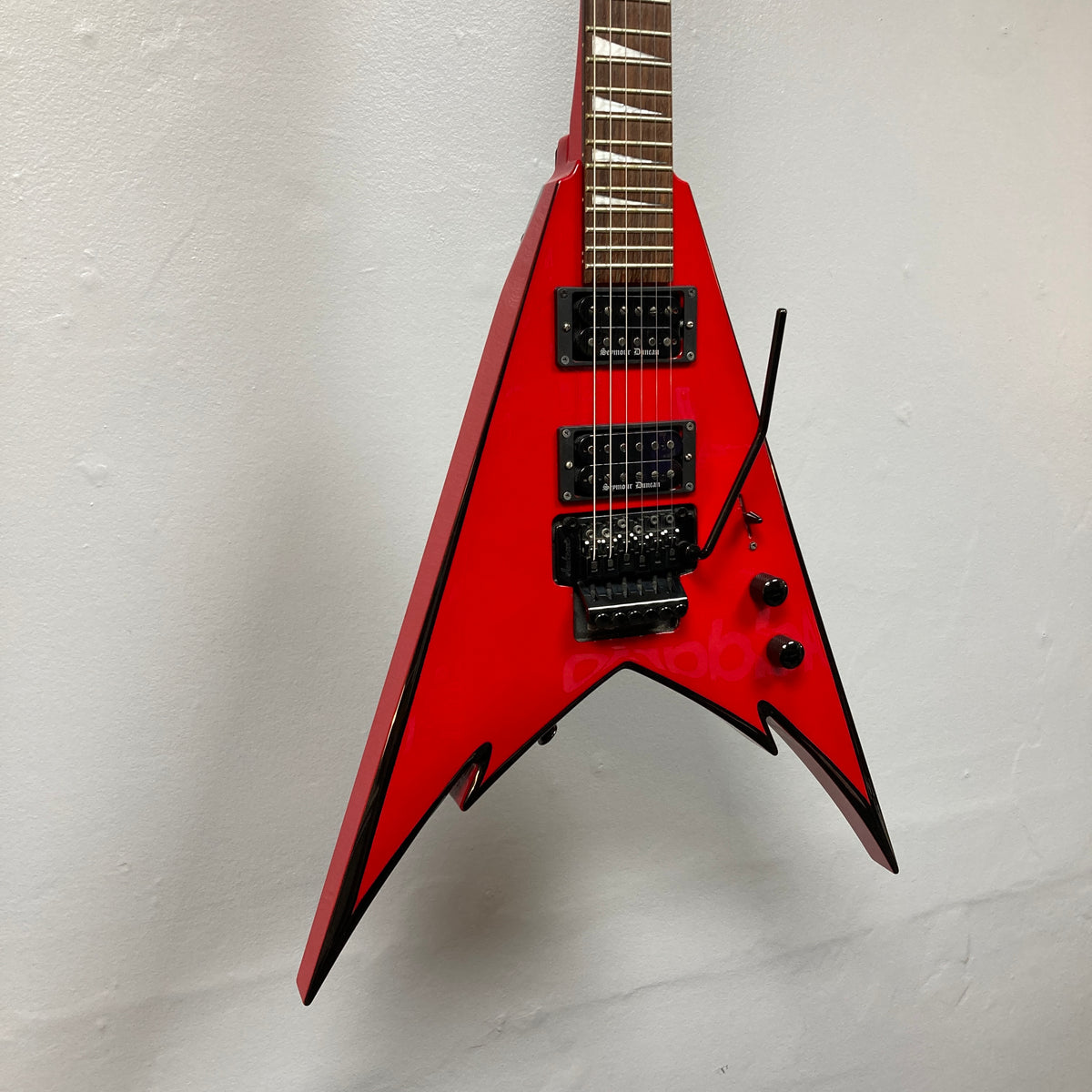 A Jackson Phil Demmel Demmelition King V electric guitar by Jackson, featuring a red pointed V-shaped body with black trim, dual humbucker pickups, and a Floyd Rose tremolo whammy bar, photographed against a plain white wall.