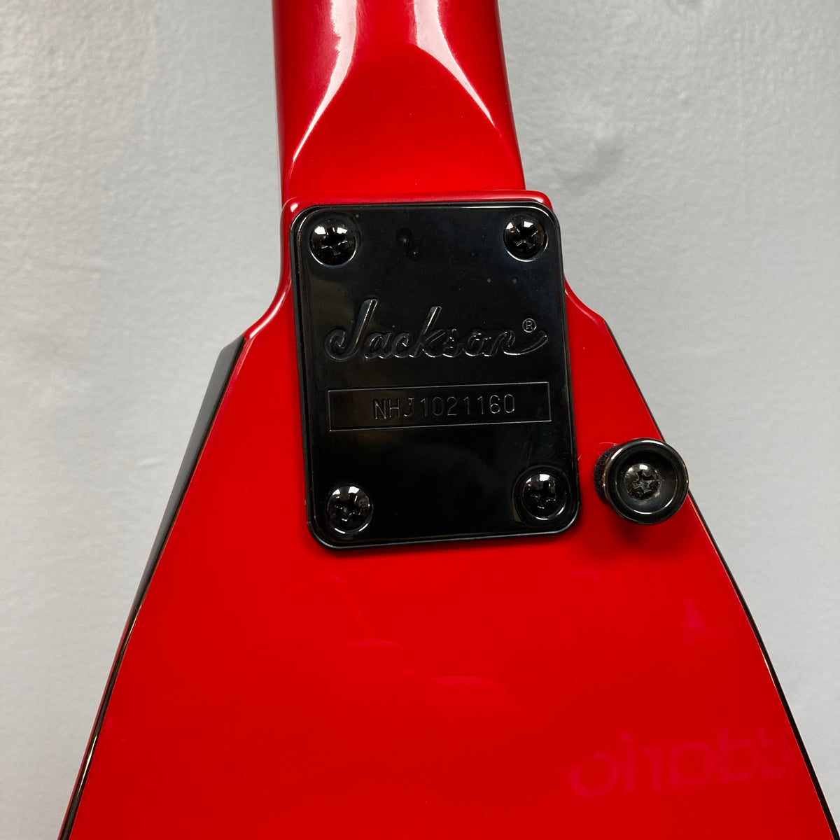 Close-up of a red Jackson Phil Demmel Demmelition King V guitar’s neck plate, showing the engraved Jackson logo and serial number NHJ021160 against a plain background. This signature model features distinctive hardware often found on high-performance instruments, such as the acclaimed Floyd Rose tremolo system.