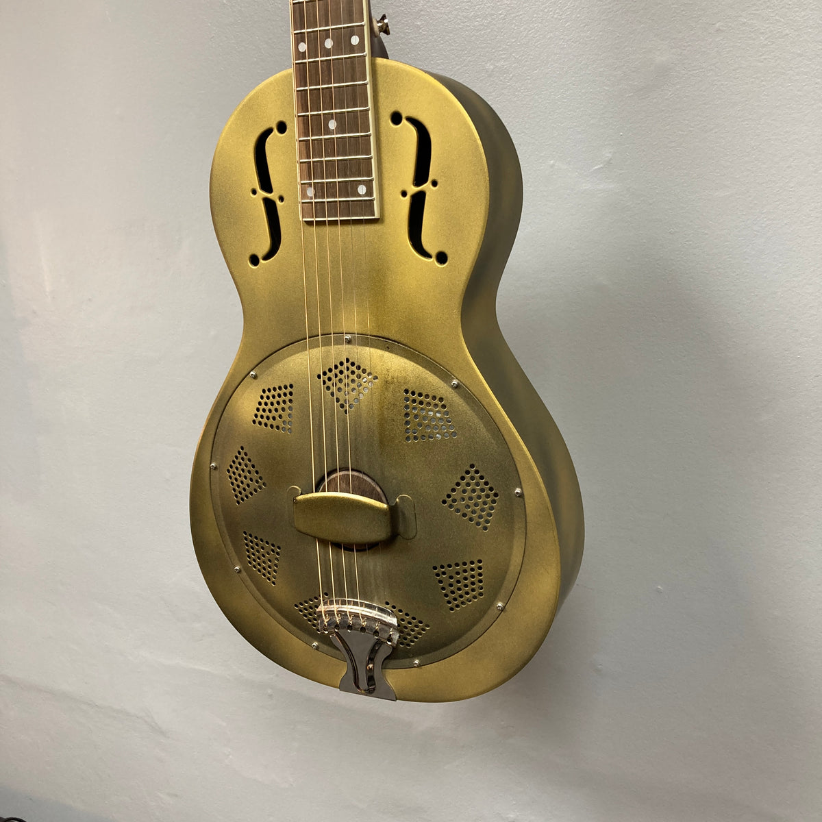 A Republic Parlor Resolian Antique Bronze Resonator guitar by Republic, featuring f-shaped sound holes and a single cone, is hanging against a plain light gray wall.