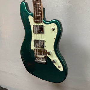 A Squier Paranormal Rascal Bass HH in Sherwood Green, featuring a white pickguard, two Fender-designed Wide Range humbucker pickups, three control knobs, and a chrome bridge, is hanging against a plain light gray wall.