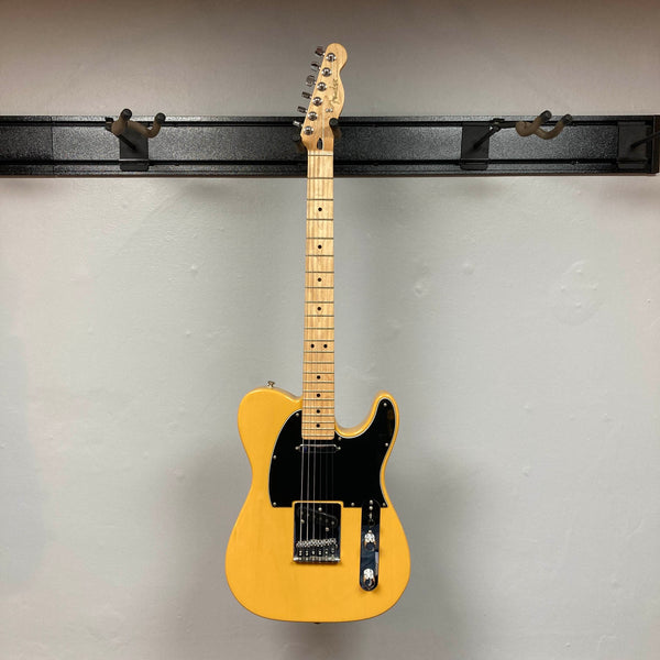 A Fender Player Telecaster 75th Anniversary Butterscotch Used electric guitar by Fender, featuring a black pickguard and maple fingerboard, is hanging against a light gray wall.