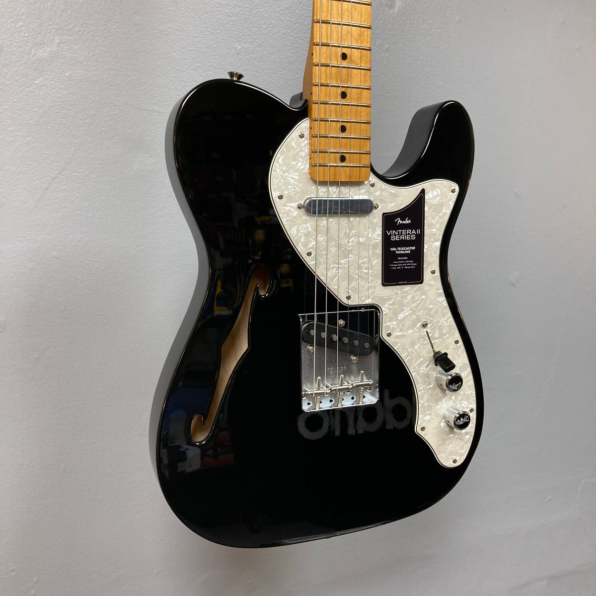 A Fender Vintera II ’60s Telecaster Thinline Black Used electric guitar by Fender, featuring a white pearl pickguard, maple neck, vintage-style Tele pickups, and an f-hole, graces the wall. Its semi-hollow ash body enhances both its classic appearance and resonant tone.