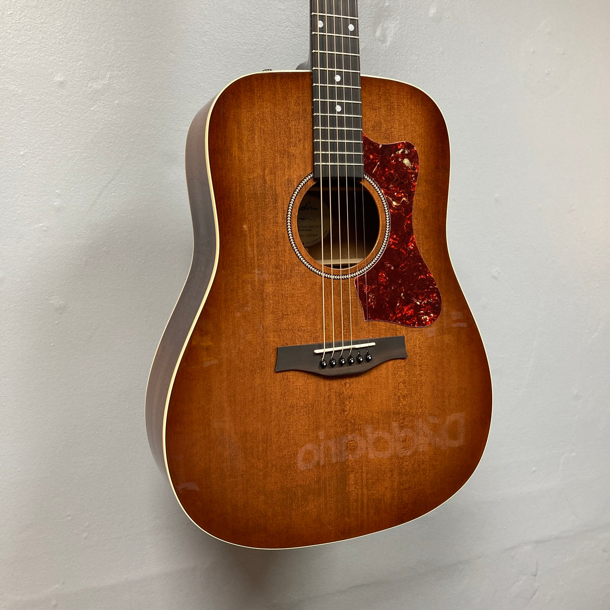 A Godin Century Maho Rustic Burst EQ SF acoustic electric guitar with a tortoiseshell pickguard is hanging on a light-colored wall.