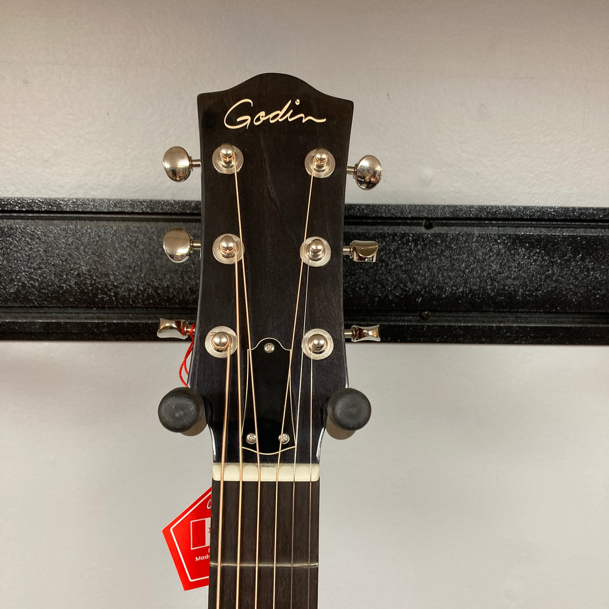 Close-up of the headstock of a Godin Century Maho Rustic Burst EQ SF acoustic guitar by Godin, featuring six tuning pegs, strings, and a red tag hanging from one peg against a plain background. This solid wood guitar blends classic design with modern versatility, making it ideal for both acoustic and electric performance.