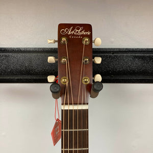 The headstock of the Art & Lutherie Roadhouse Denim Blue Q-Discrete acoustic guitar features tuning pegs, strings, and a red tag, shown against a white wall and black rack.