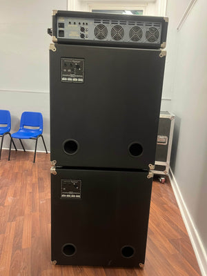 An Ashdown UK Built ABM Bass Rig Full Stack with EVO II 900 Head, 410T and 115-500 cabinets in flight cases stands against a wall on wood flooring; two blue chairs are to the left.