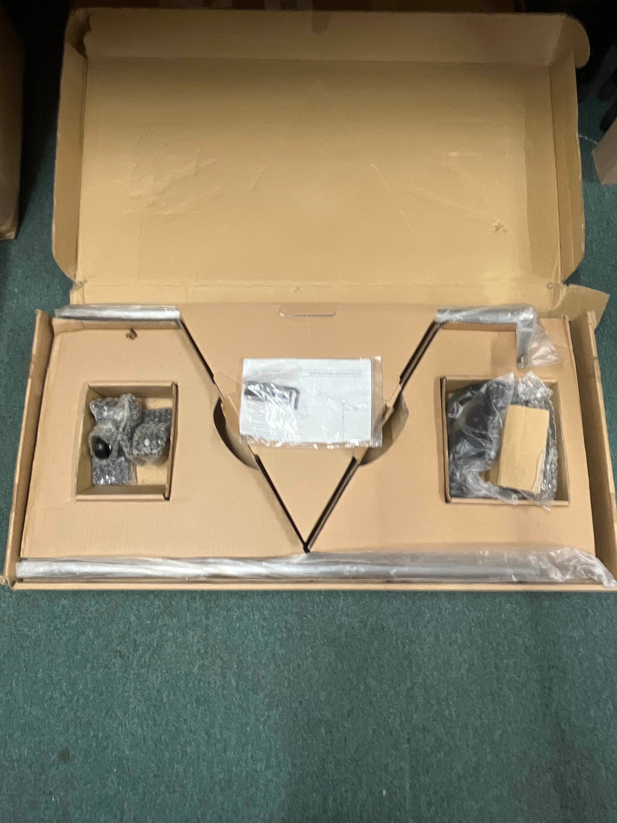 Opened cardboard box containing disassembled parts of an Atlas Sound SB36W Professional Studio Boom Microphone Stand, some wrapped in plastic and bubble wrap, placed on a green carpet.