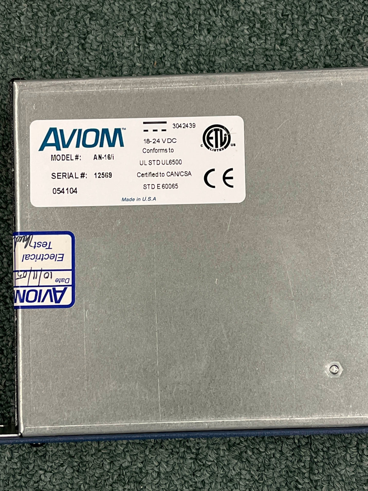 An Aviom AN-16/i A-Net Input Module by Aviom, featuring certification marks, serial number 12569, and voltage specs, is shown on a green textured surface—perfect for professional live sound setups.