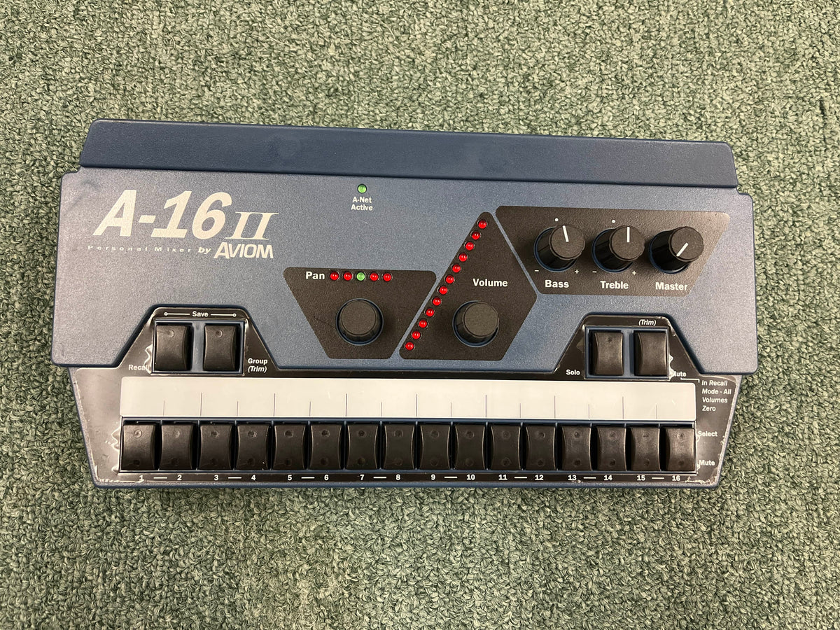 The Aviom AN-16/II A-Net Input Module by Aviom, shown on a green carpet, features labeled controls for pan, volume, bass, treble, master, and 16 channel buttons—perfect for personal mixing networks.