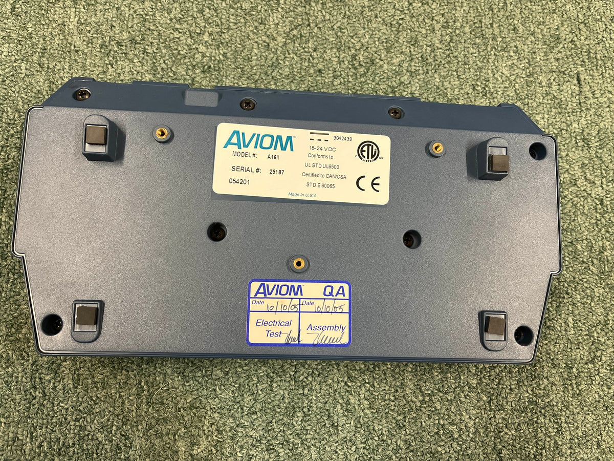 The underside of an Aviom AN-16/II A-Net Input Module shows model and serial number labels, certification marks, and quality assurance stickers on a green carpet.