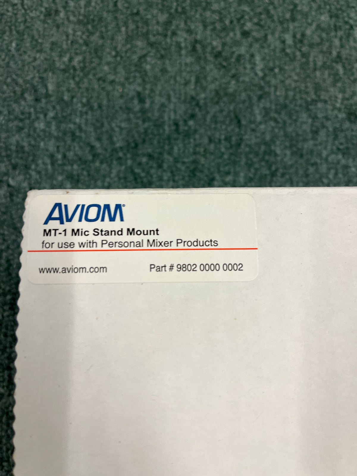 A white label on the box says "Aviom MT-1 Mic Stand Mount" by Aviom, lists a part number and website, and rests on a green textured background.