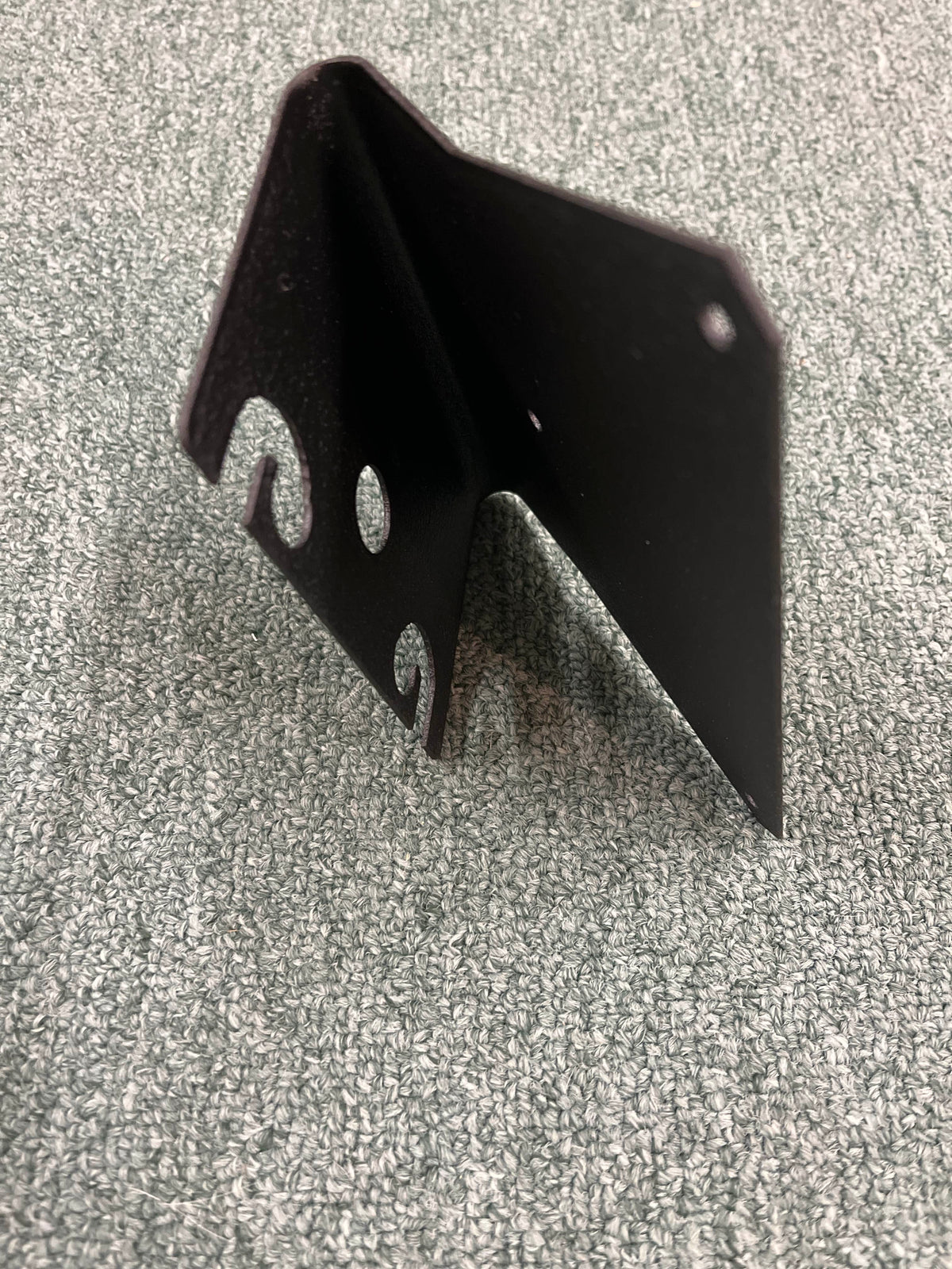 The Aviom MT-1 Mic Stand Mount, a black metal bracket with multiple holes and notches by Aviom, stands upright on a textured gray surface.