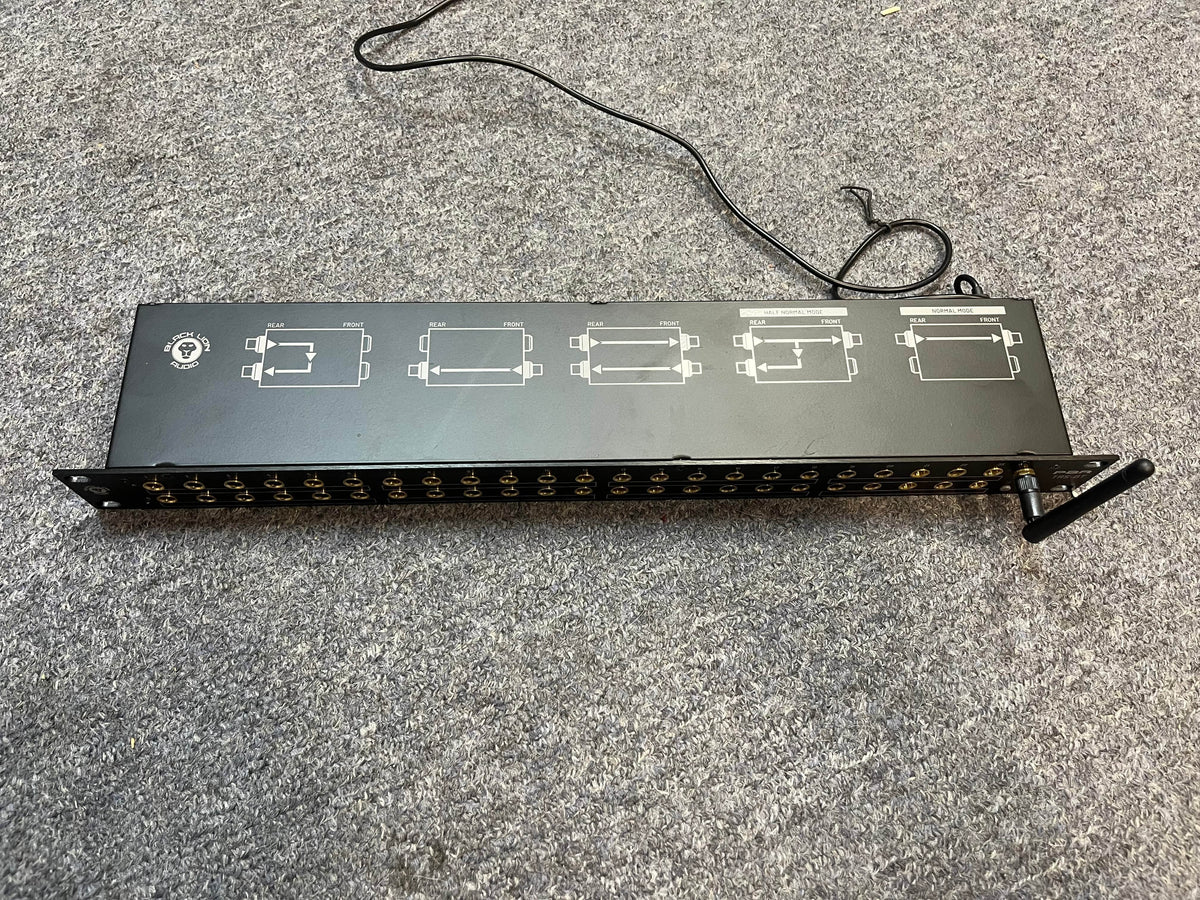 A Black Lion Audio PBR TRS-BT 46-Point Gold-Plated TRS Patchbay with Bluetooth (1 RU) featuring front ports, connectors, a thin cable, and white diagrams on top sits on a gray carpet. This device also serves as a Bluetooth receiver.