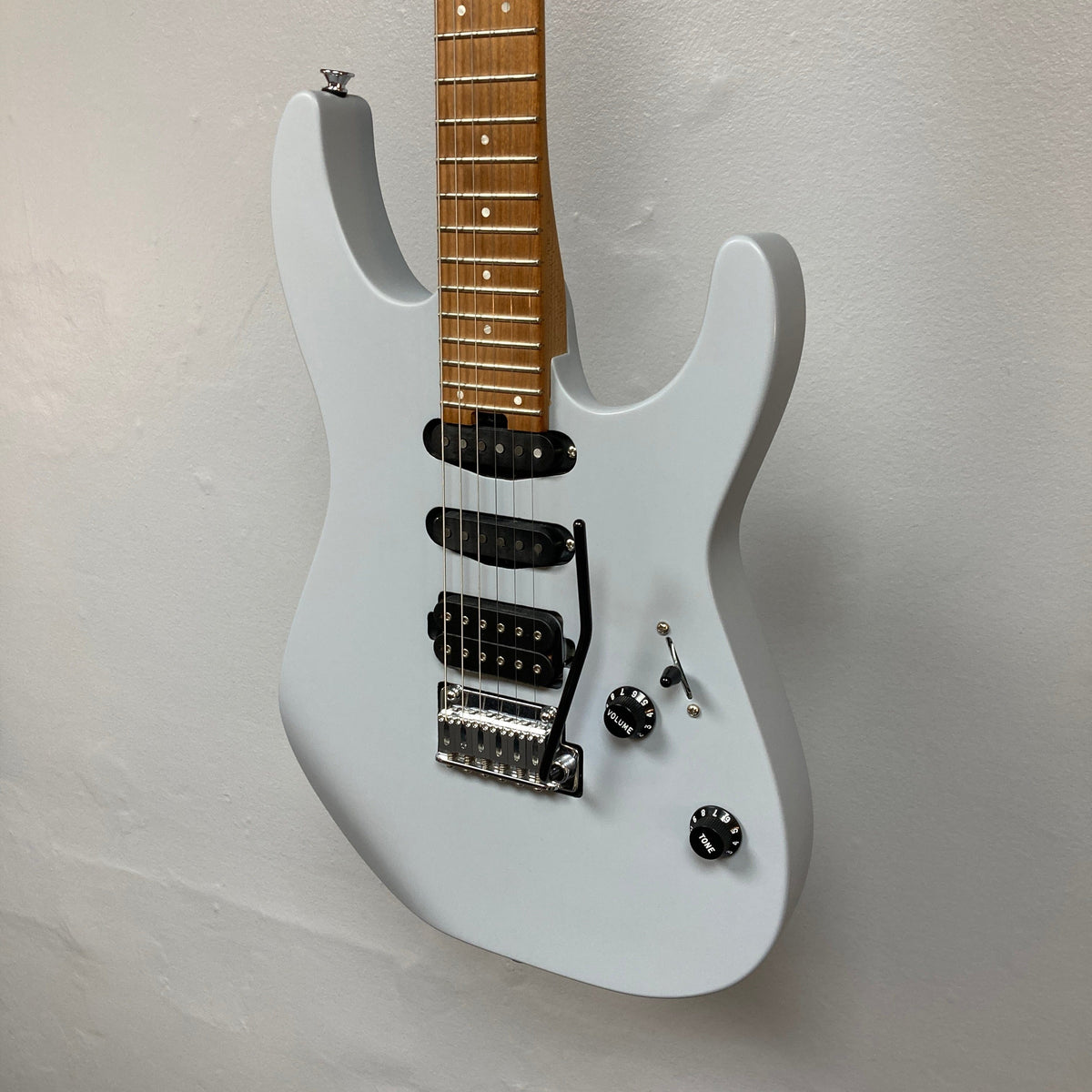A Used Charvel DK24 HSS 2PT CM Electric Guitar in gray with a maple neck, rosewood fretboard, Seymour Duncan pickups, volume and tone knobs, and a tremolo bridge by Charvel hangs on a white wall.