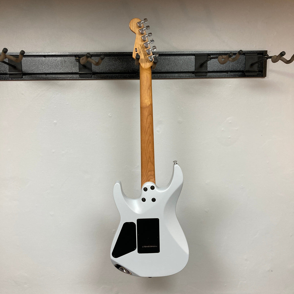 A Used Charvel DK24 HSS 2PT CM Electric Guitar - Gray by Charvel hangs by its neck on a wall-mounted metal rack. The back view shows the wooden neck and black access panels against a plain white wall.