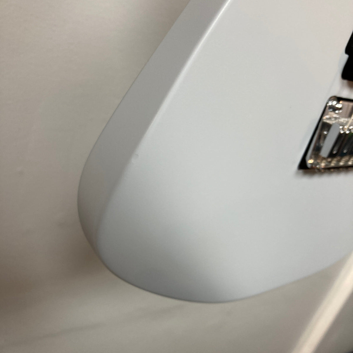 Close-up of the bottom edge of a gray Used Charvel DK24 HSS 2PT CM Electric Guitar body against a white wall, with chrome bridge hardware partially visible on the right.