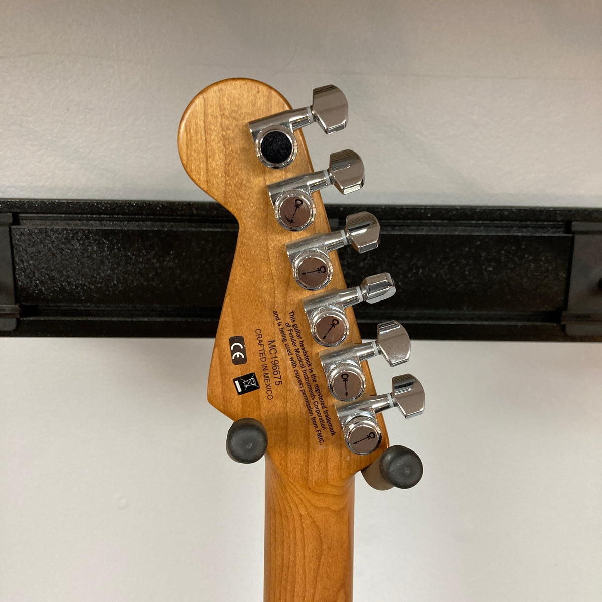 The image shows the back of a Charvel DK24 HSS 2PT CM Electric Guitar - Gray headstock with six chrome tuners, brand and serial labels on the wood, resting on a black hanger against a white wall.