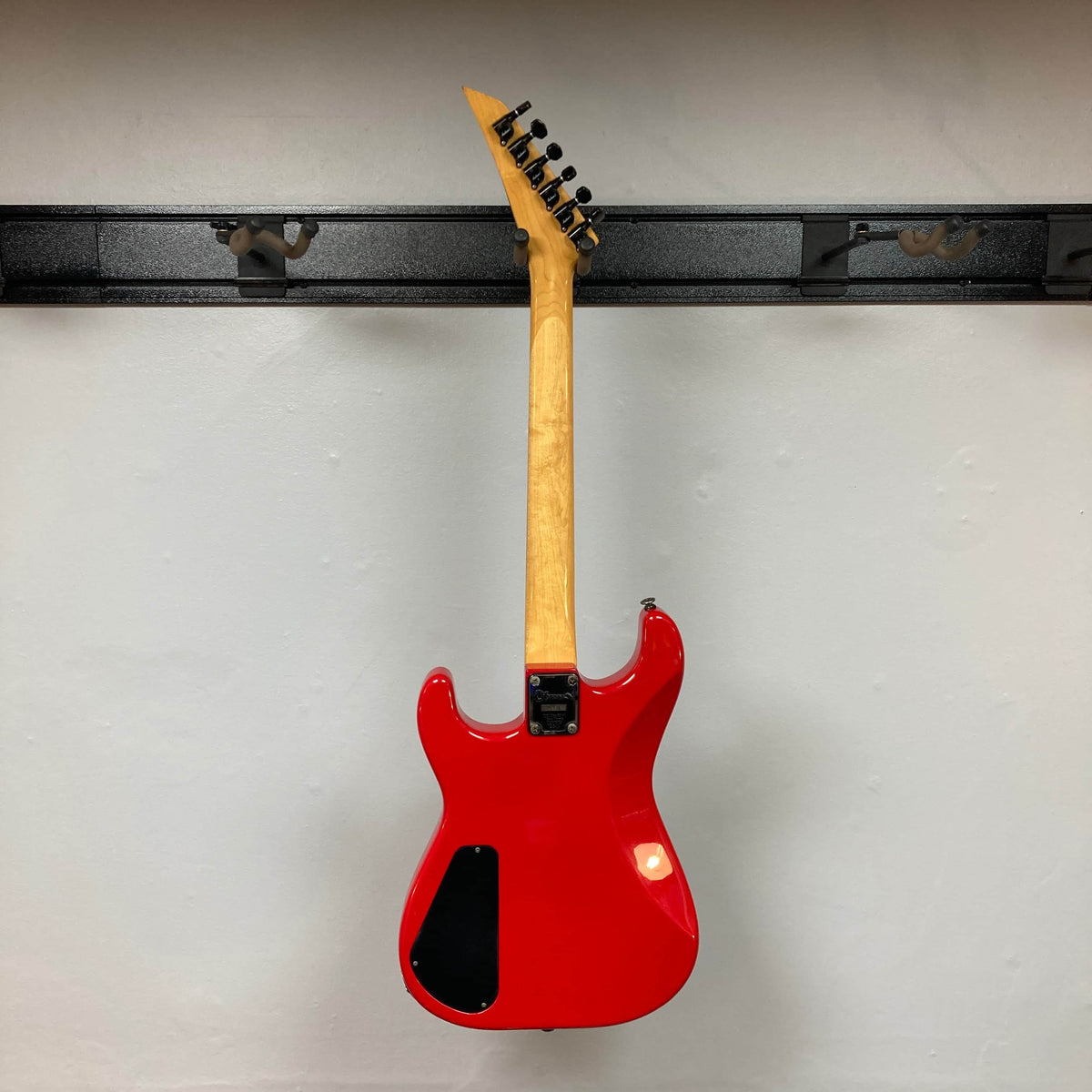 A Charvel Model 4 HSS Red w/OHSC Consign electric guitar by Charvel hangs on a black wall mount, photographed from the back to showcase its neck and body.