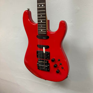 A Charvel Model 4 HSS Red w/OHSC Consign electric guitar with a black bridge, multiple control knobs, and three pickups in HSS configuration hangs on a white wall.