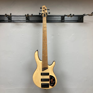 Cort Artisan B5 Element Natural Used bass guitar mounted on a wall, showcasing its ergonomic design and premium components like the MetalCraft M5 bridges.