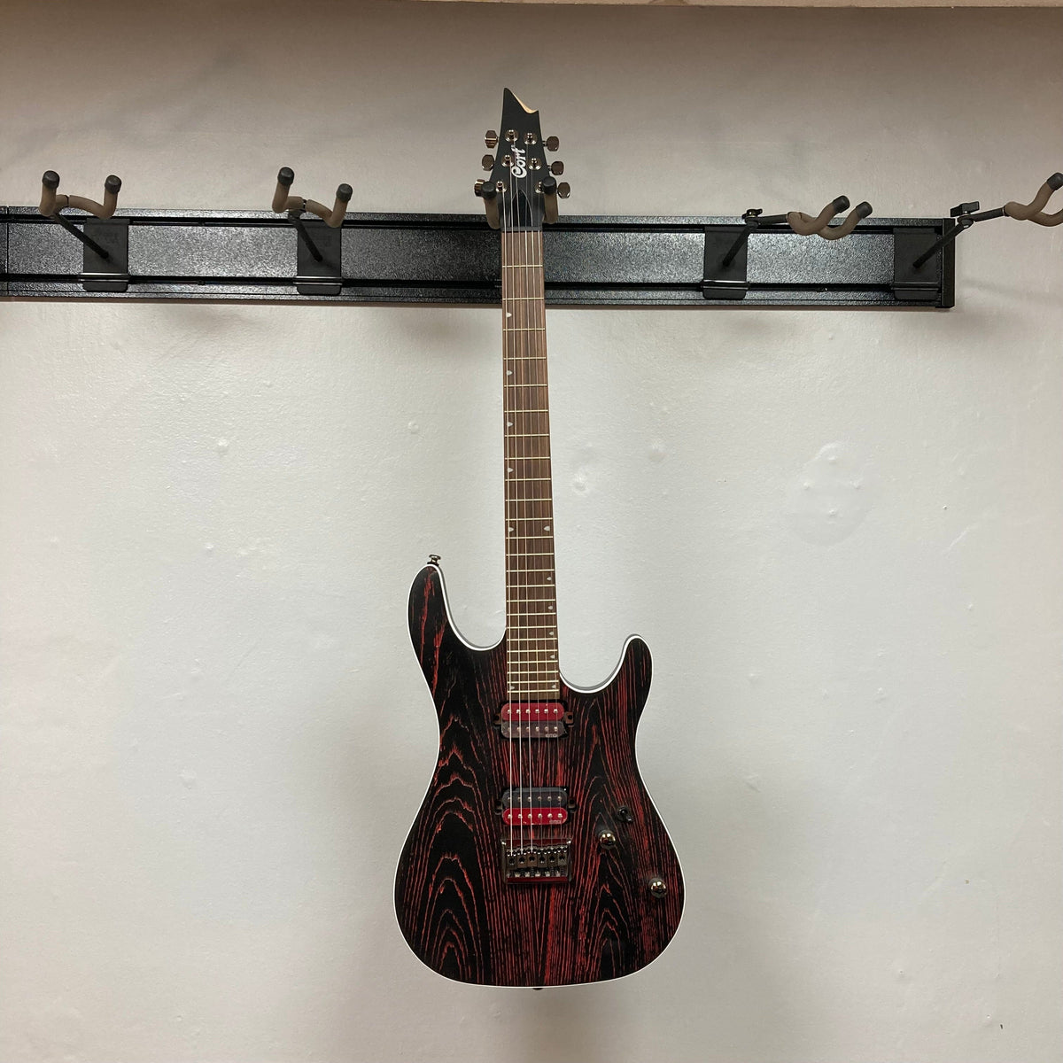 Cort KX300 EBR Etched Black Red electric guitar with ash top and EMG® RetroActive Super77 humbuckers, displayed on a wall.