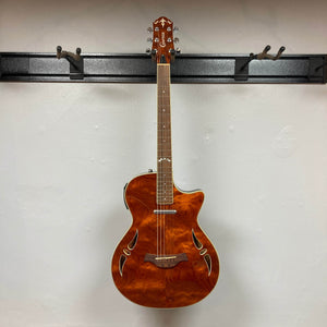 A used Crafter SA-BUB Acoustic Electric Guitar with a glossy figured wood top, cutaway body, decorative sound holes, and six strings hangs on a wall-mounted black rack against a white wall—ideal for electric-acoustic performances.