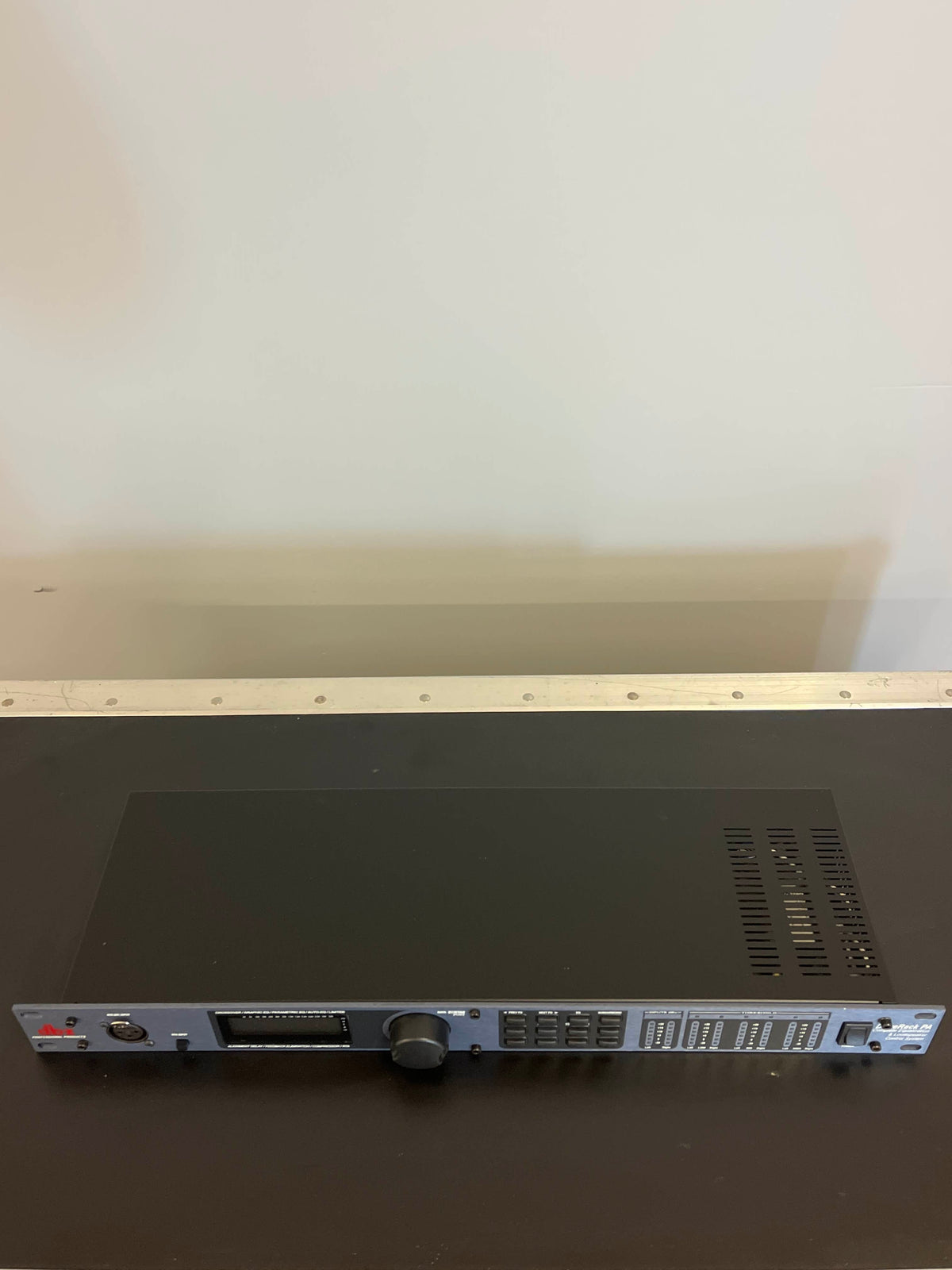 A dbx DriveRack PA Complete Loudspeaker Management System in black, featuring ventilation slots, buttons, and knobs, rests on a dark surface against a plain light-colored wall.