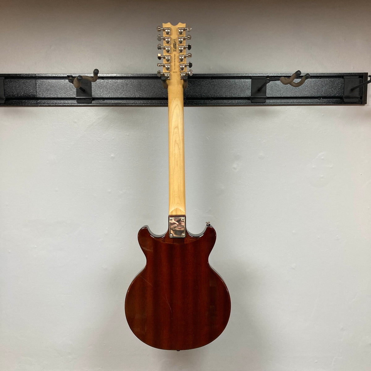 A Dean Boca Trans Amber Burst 12 String electric guitar by Dean hangs on a wall-mounted rack, displayed from the back to show its natural wood neck and dark amber burst finish, highlighting its unique twelve-string setup.