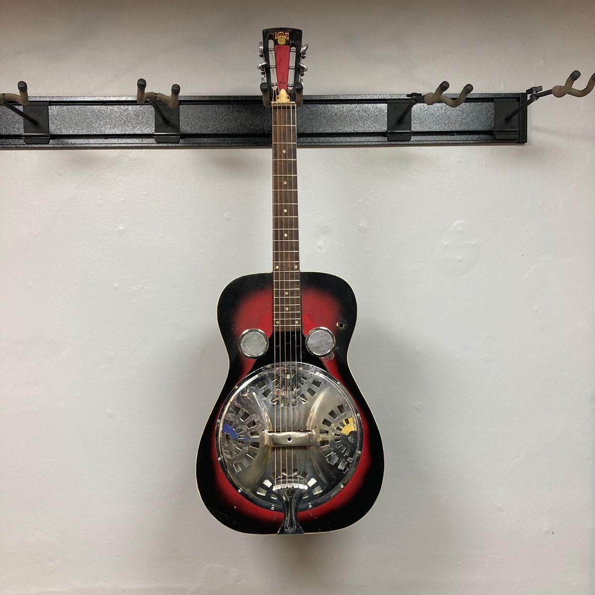 Dobro Model 60 DS Round Neck 1970s Red Burst resonator guitar hanging on a wall, showcasing its well-used condition with visible blemishes.