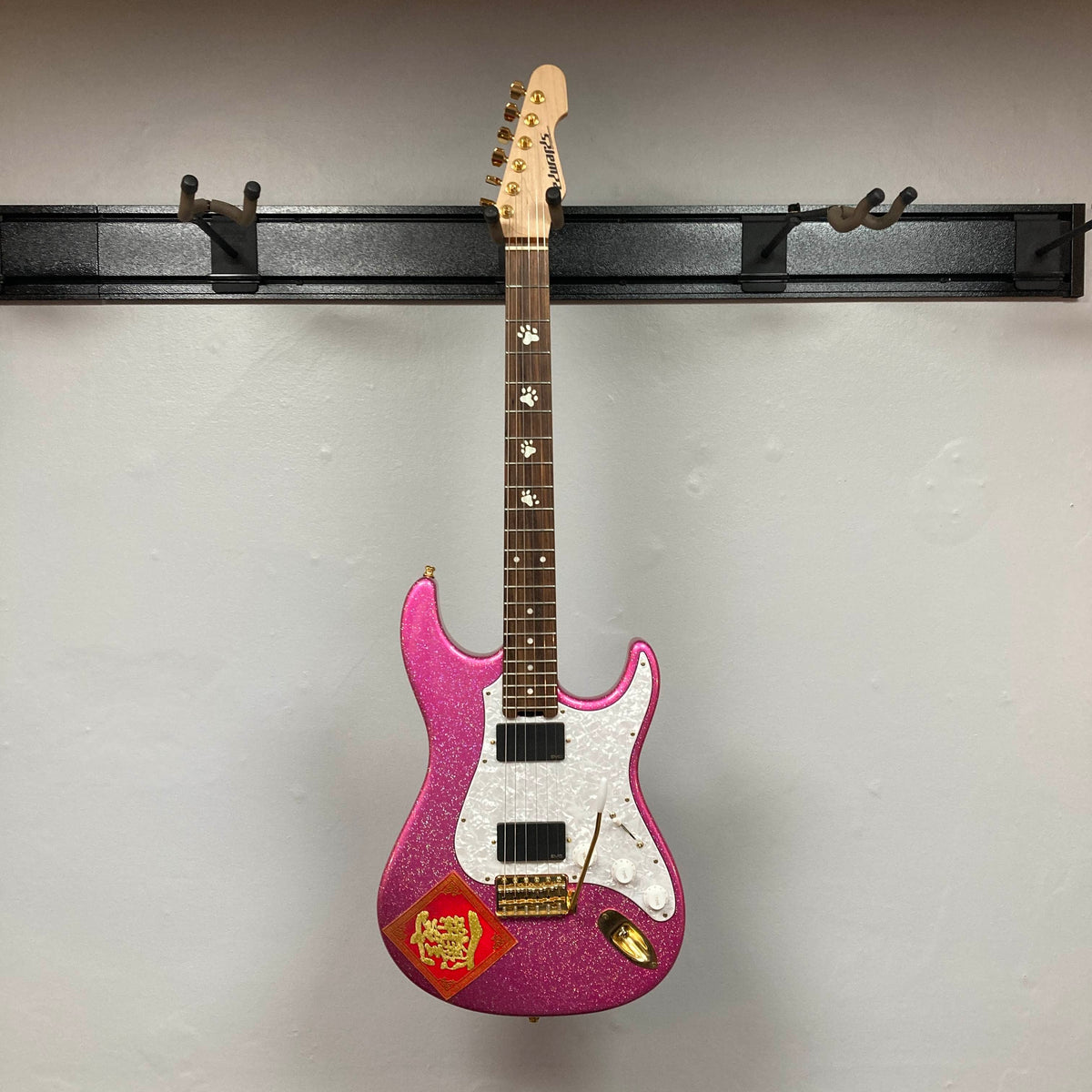 An Edwards ESP E-SN-185-TO Takayoshi Ohmura electric guitar in Twinkle Pink with a white pickguard and a red sticker is hanging on a black wall mount against a plain light-colored wall.