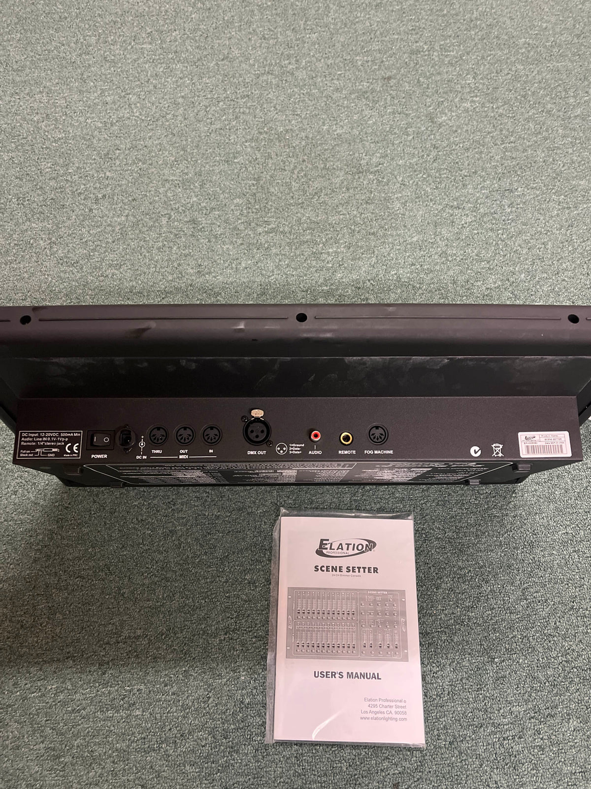 Back panel of the Elation Scene Setter 24-Channel DMX Dimmer Console by Elation, displaying multiple input/output ports, shown above its user manual on a carpeted floor—ideal for professional lighting control and scene programming.