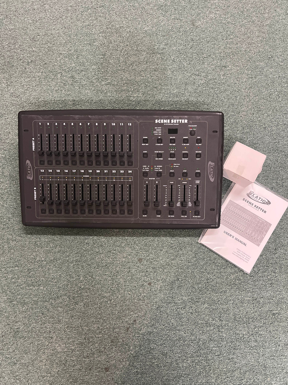 An Elation Scene Setter 24-Channel DMX Dimmer Console by Elation rests on a gray carpet, accompanied by its user manual and warranty card.