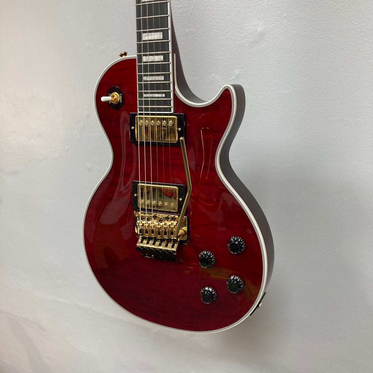Epiphone Alex Lifeson Les Paul Custom Axcess, red electric guitar with gold hardware, displayed on a wall, featuring Floyd Rose tremolo and Epiphone pickups.