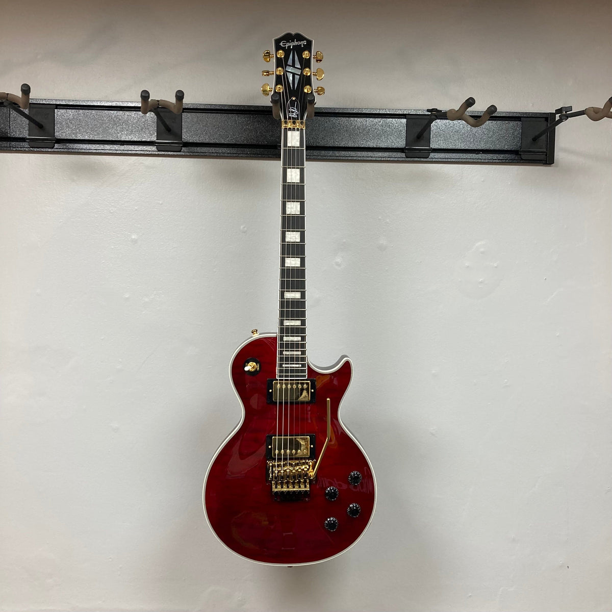 Epiphone Alex Lifeson Les Paul Custom Axcess w/Case, featuring a red body with gold hardware, displayed on a wall hook.