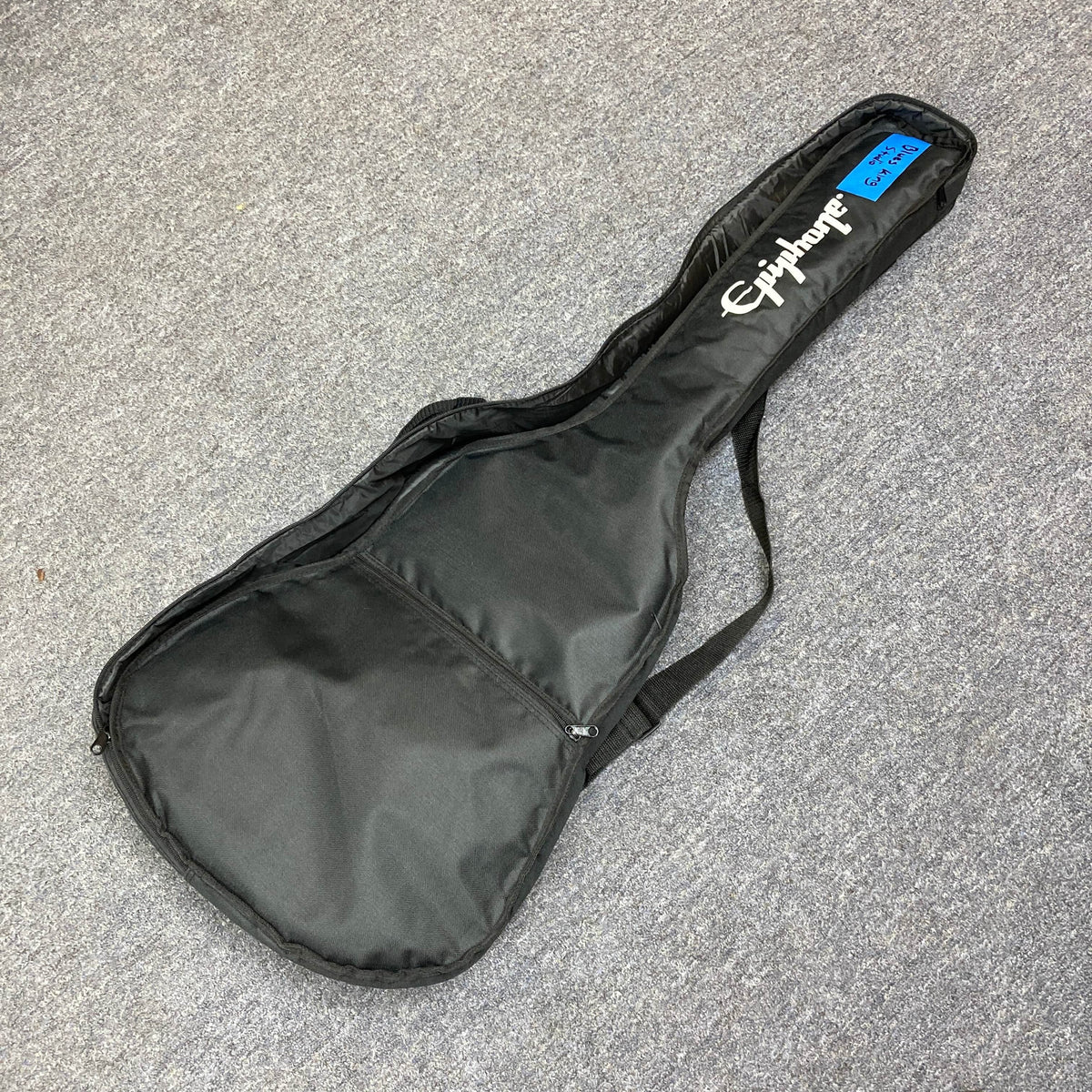 A black Epiphone soft case, ideal for the Epiphone Blues King Studio Dark Burst acoustic guitar, features a shoulder strap and front zipper pocket. It rests on gray carpet with a blue numbered sticker on the headstock area.