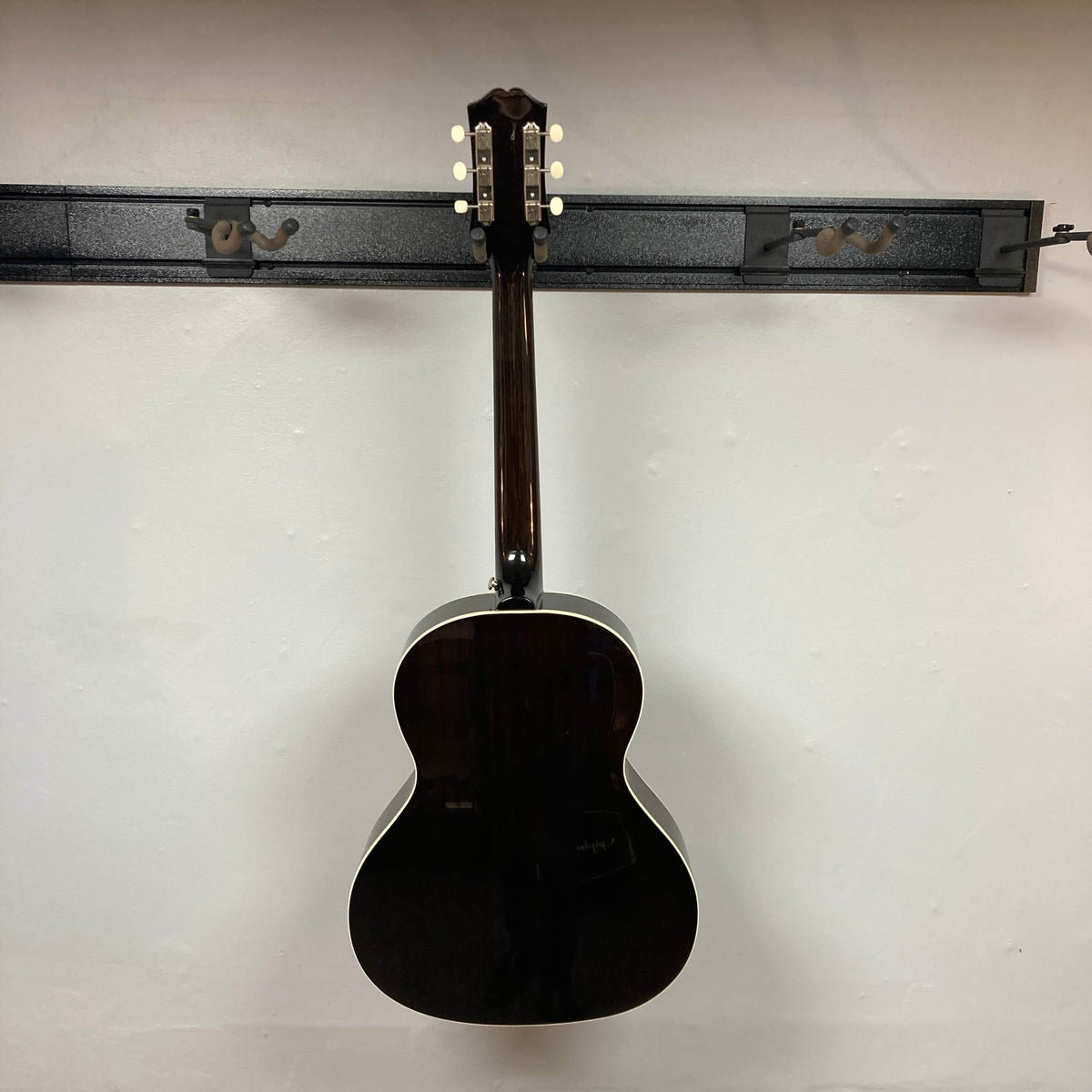 An Epiphone Blues King Studio Dark Burst acoustic guitar by Epiphone hangs on a black wall rack. Displayed from the back, it features a dark brown finish with white trim and visible tuning pegs on the headstock.