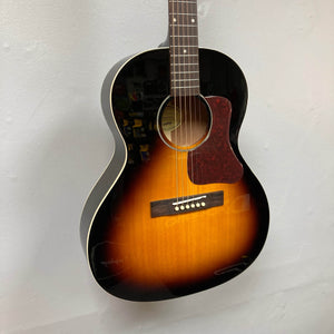 An Epiphone Blues King Studio Dark Burst acoustic guitar by Epiphone hangs on a white wall, featuring steel strings, a reddish-brown pickguard, wooden bridge, and sunburst finish inspired by classic Gibson designs.