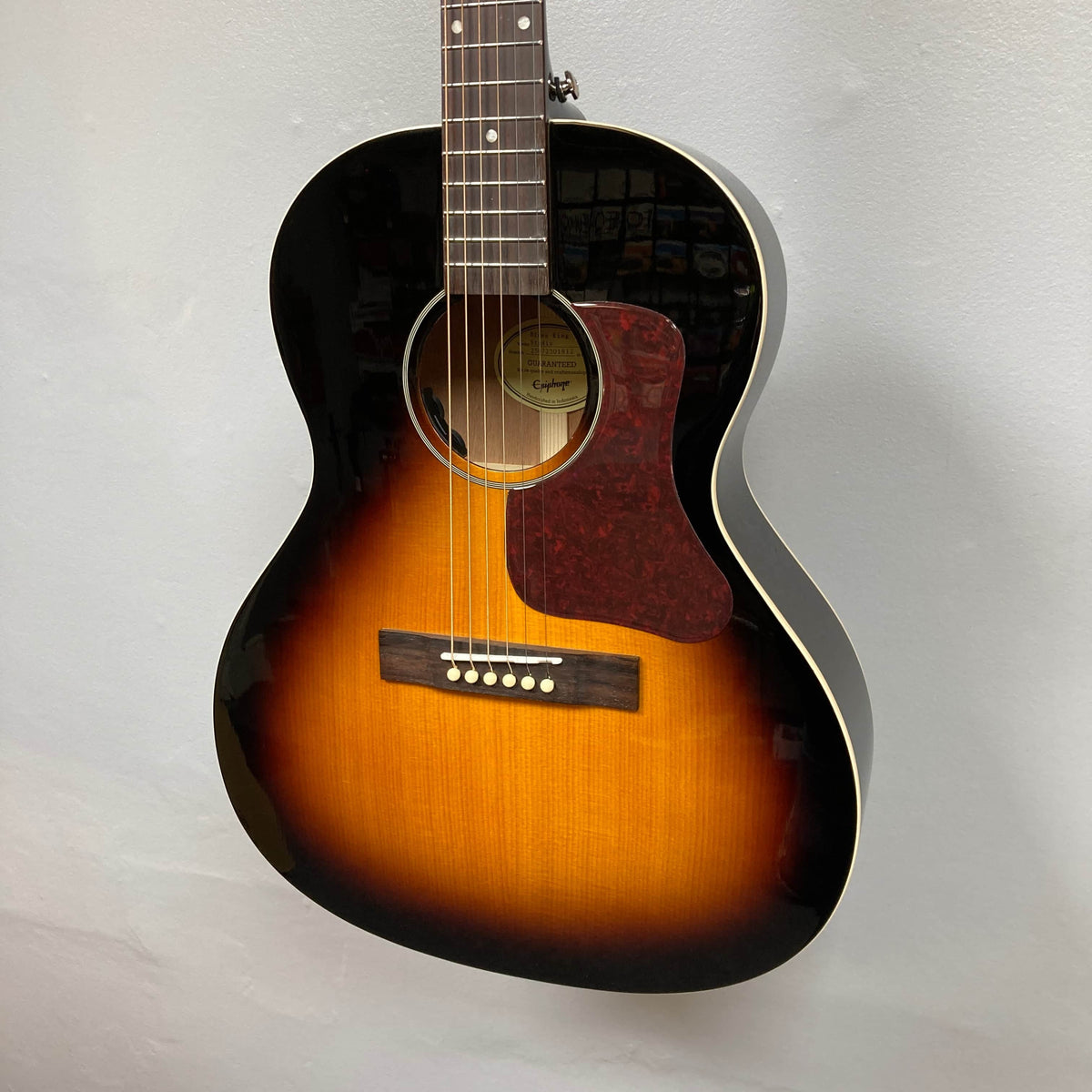 An Epiphone Blues King Studio Dark Burst parlor guitar by Epiphone, featuring a dark outer edge, yellow-orange center, reddish pickguard, and six strings, hangs against a plain light-colored wall.