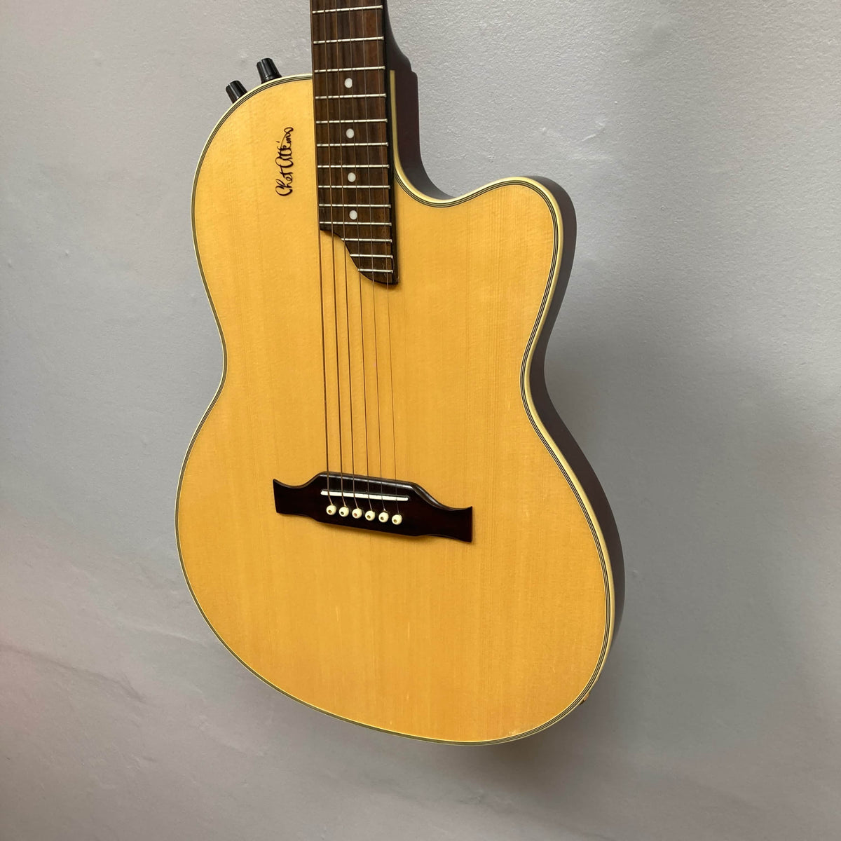 An Epiphone Chet Atkins SST Studio Natural acoustic-electric guitar, made in Korea, is displayed on a light gray wall. It features six tuning pegs, control knobs, a cutaway body, and comes with a hard case.