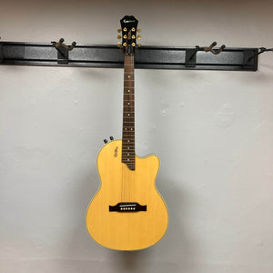 An Epiphone Chet Atkins SST Studio Natural (Korea) acoustic-electric guitar, known for its feedback resistance, hangs on a black wall rack against a white wall. This used consignment includes a hard case and features gold tuners and a piezo pickup.