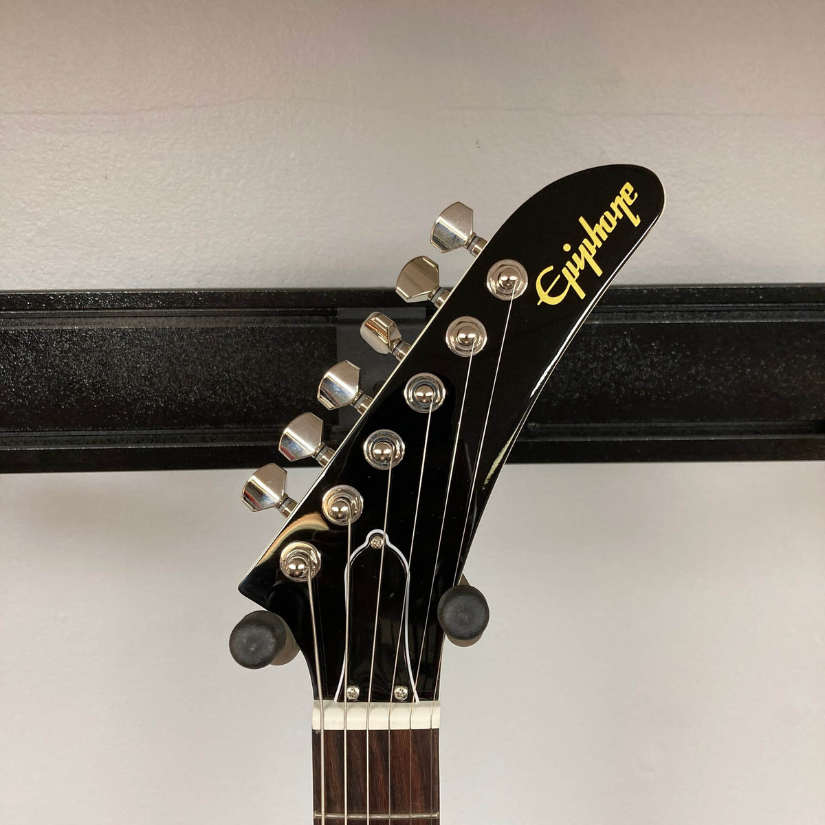 Close-up of the headstock of the Epiphone Explorer 80s EMG Classic White guitar by Epiphone, showing six tuning pegs against a light wall and a horizontal black metal bar.