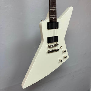 A classic white Epiphone Explorer 80s EMG electric guitar with an angular body, dual humbuckers, four control knobs, and a metallic bridge hangs on a plain light gray wall. A gigbag is included.