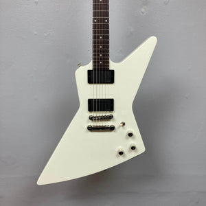 The Epiphone Explorer 80s EMG Classic White w/Gigbag features a modern angular body, two black pickups, three control knobs, and a toggle switch. Displayed against a plain gray wall. Brand: Epiphone.