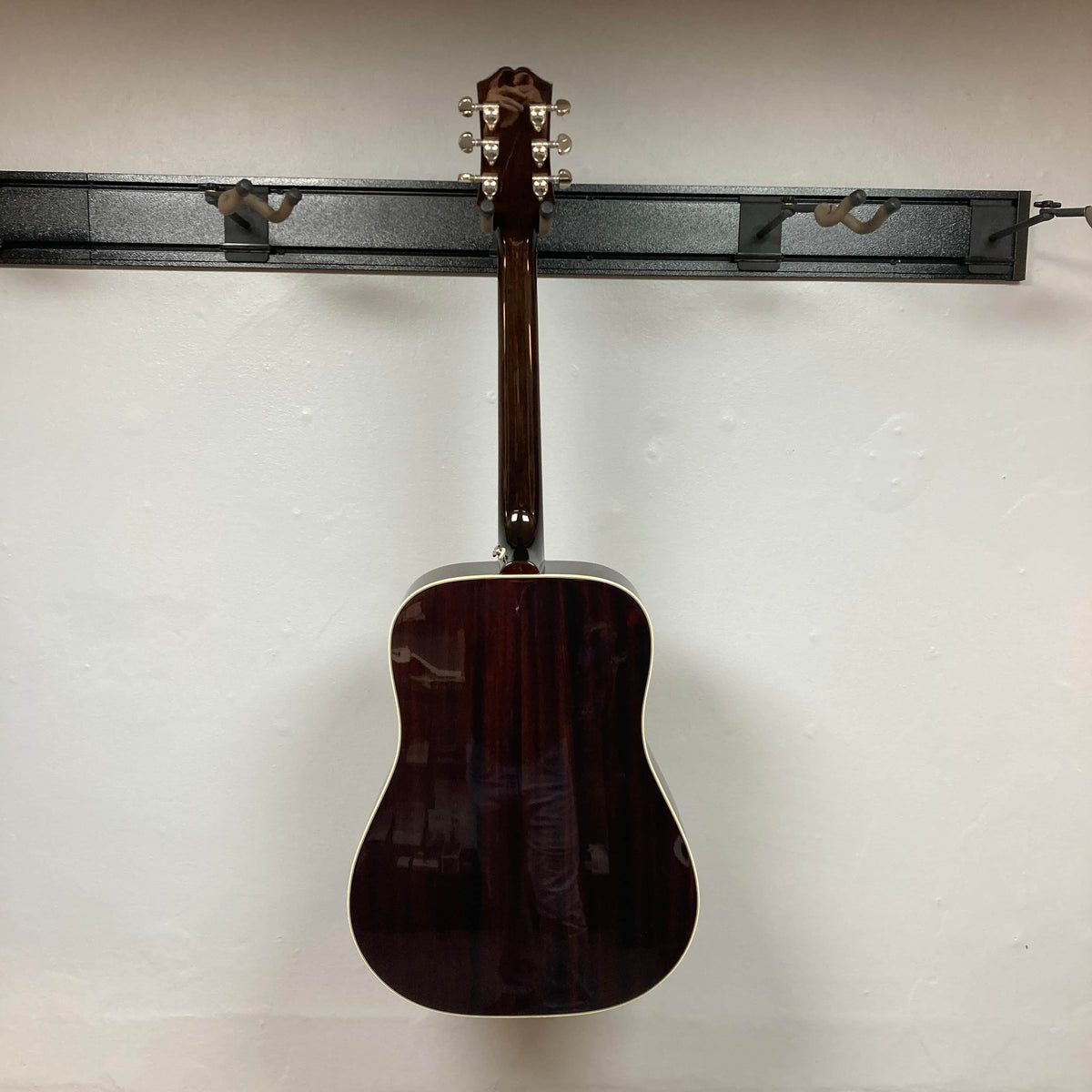 An Epiphone Hummingbird Studio Antique Natural acoustic guitar with a solid spruce top is displayed from the back, hanging centrally on a black wall rack against a plain white background. Includes gigbag.