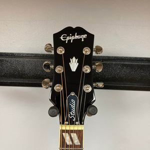 Close-up of the Epiphone Hummingbird Studio Antique Natural headstock, showing six tuning pegs, the Epiphone logo, emblem, and a "Studio" truss rod cover—part of this model with a solid spruce top. Comes with gigbag.