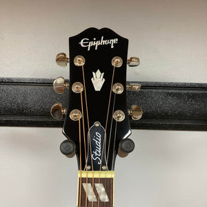 Close-up of the headstock of the Epiphone Hummingbird Studio Faded Cherry Sunburst acoustic-electric guitar, featuring gold tuners, Epiphone logo, decorative inlay, and "Studio" truss rod cover against a plain light wall.